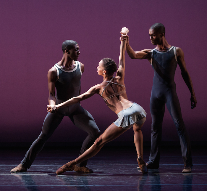Two men hold a woman's wrists as she balances precariously on pointe while her pelvis tilts behind her. 